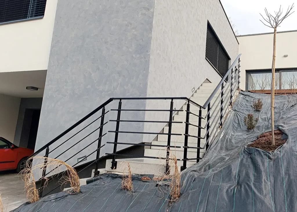 Černé kovové zábradlí s vodorovnými profily na venkovním schodišti u moderního domu.