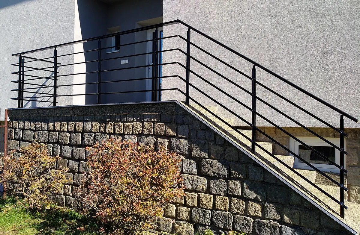 Černé kovové zábradlí s vodorovnými pruty na venkovním schodišti u domu.